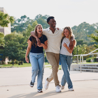 Student has his arms around two other students, walking on campus and laughing