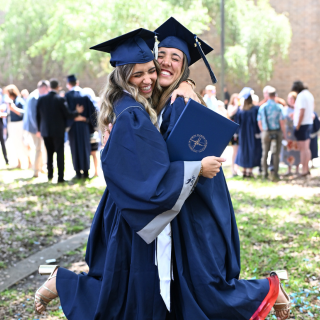 Two girls in blue graduation regalia smile and hug