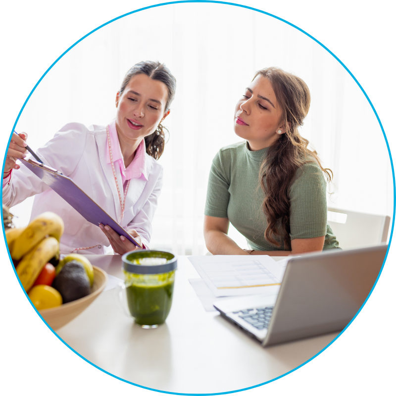 Two females, one dietitian, one dietetic student siting at a table with a clipboard, computer, papers, a cup of green juice and a bowl of fruit. 