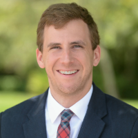 Headshot of Caleb Garrett smiling, in a sunny day with trees and garden as background.