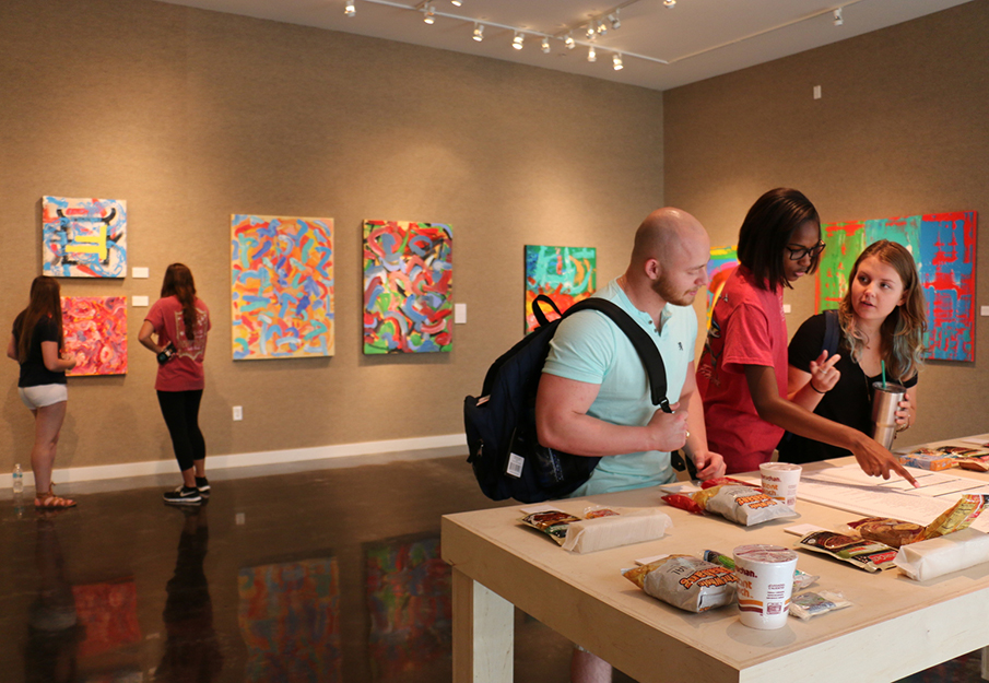 A vibrant art gallery showcasing colorful abstract paintings with visitors engaging in discussion at a central table.