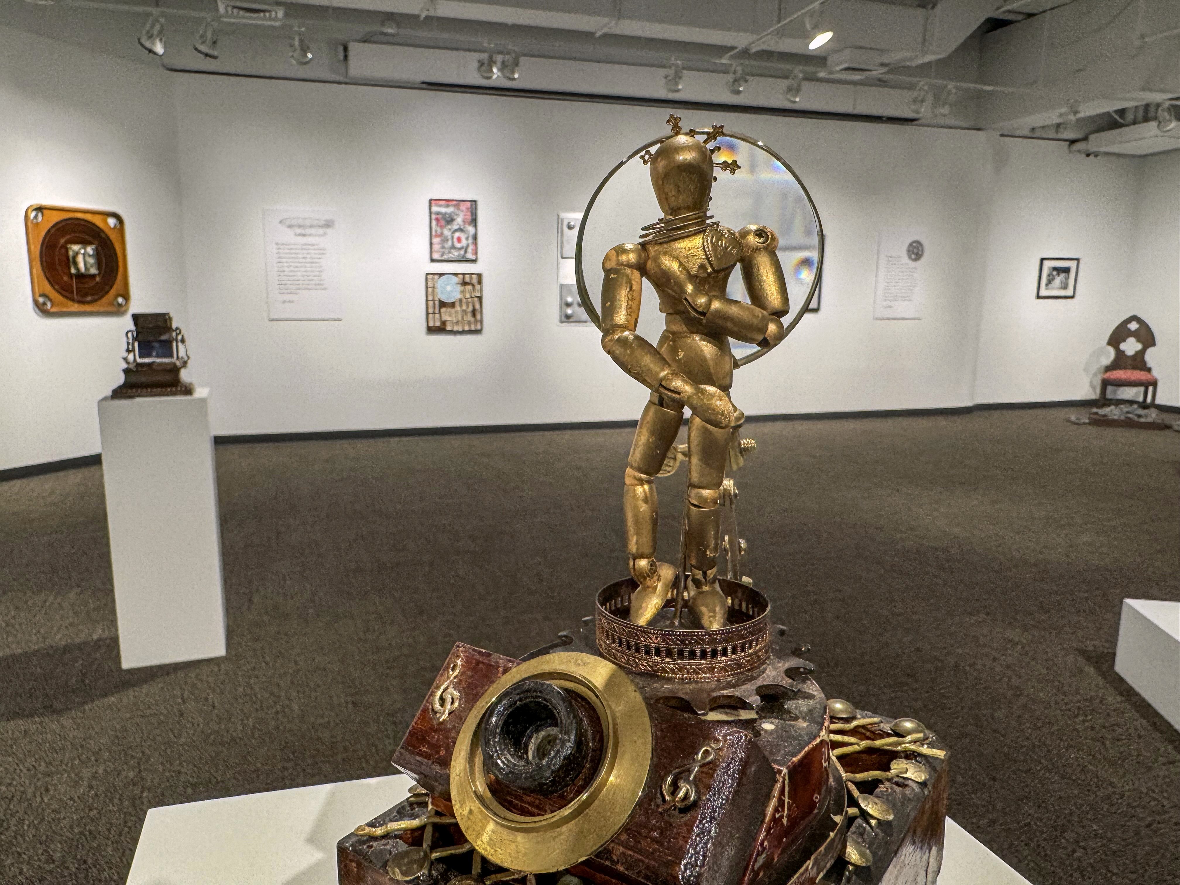 A golden robot statue stands atop a display base in an art gallery, with various artworks visible in the background.