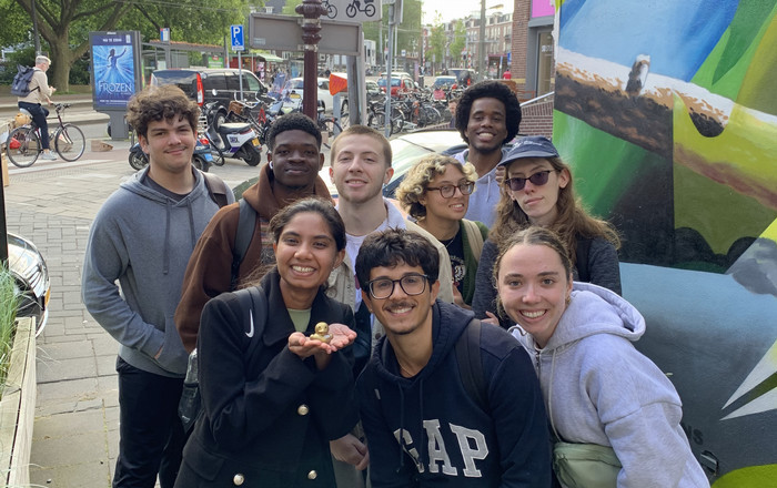 Nine students pose with a mini rubber duckie in front of a multicolored mural with bicycles in the background.