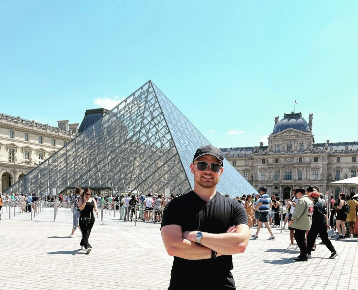 A male student wearing a black t-shirt and baseball cap poses in front of the Louvre (a large glass pyramid) in a crowded area of Paris