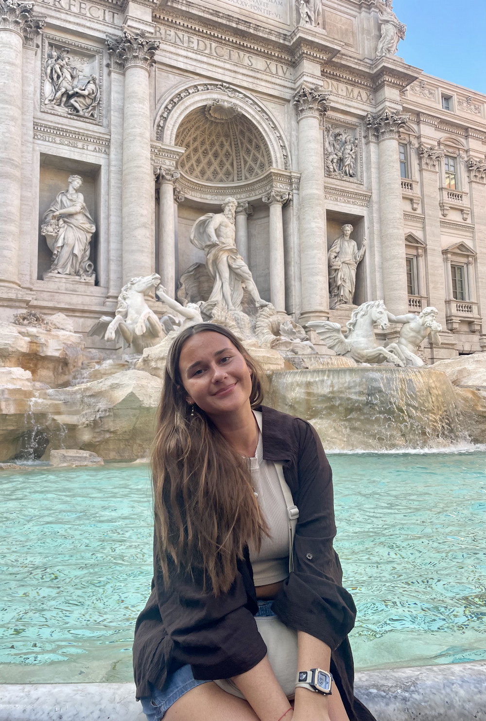 A smiling female student sits in front of a large marble fountain with elaborate sculptures and bright blue water in Italy.