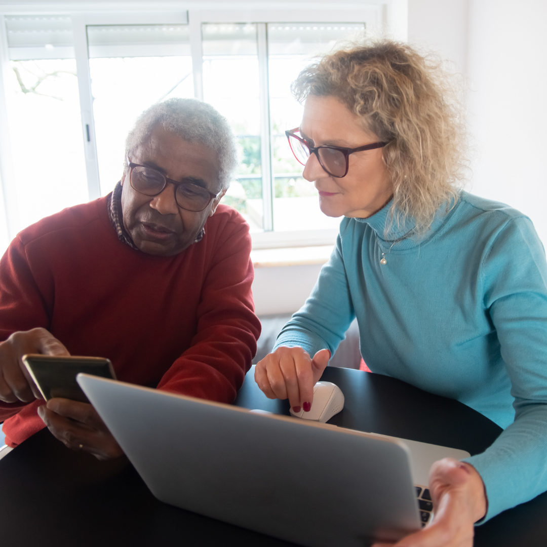two individuals sitting at desk with laptop and looking at a phone screen