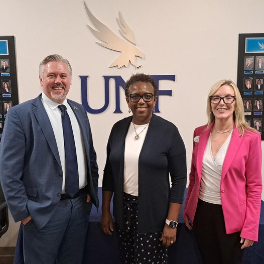 Colin, Almeta, and Kathleen smiling for a photo behind a UNF sign